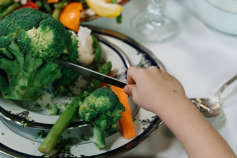 A child uses a knife to cut fresh broccoli and vegetables on a plate. Healthy eating concept.