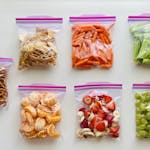 An assortment of fruits, vegetables, and snacks neatly packed in zip lock bags on a white background.