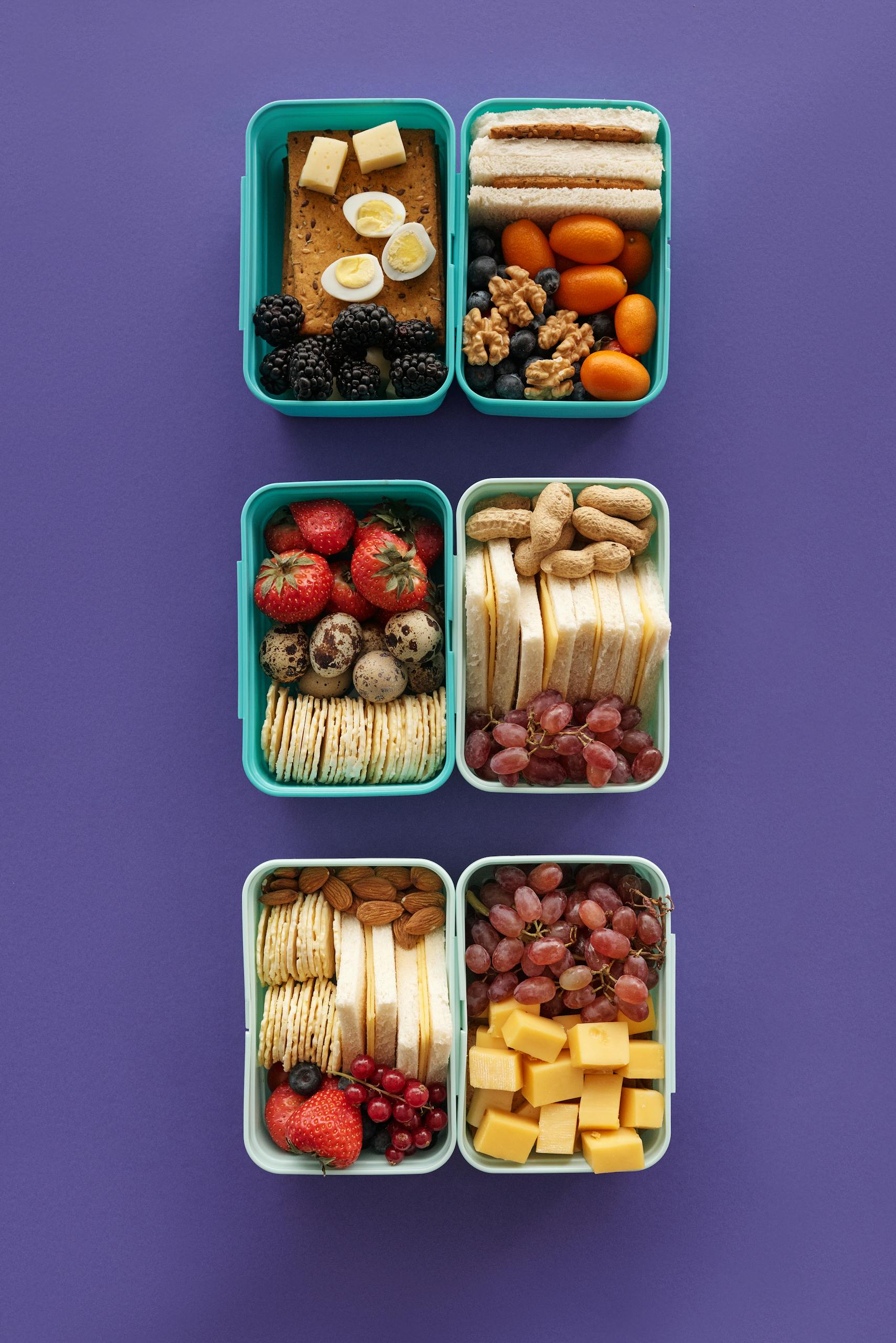 A vibrant flat lay of assorted lunch boxes with fruits, nuts, and cheese on a purple background.