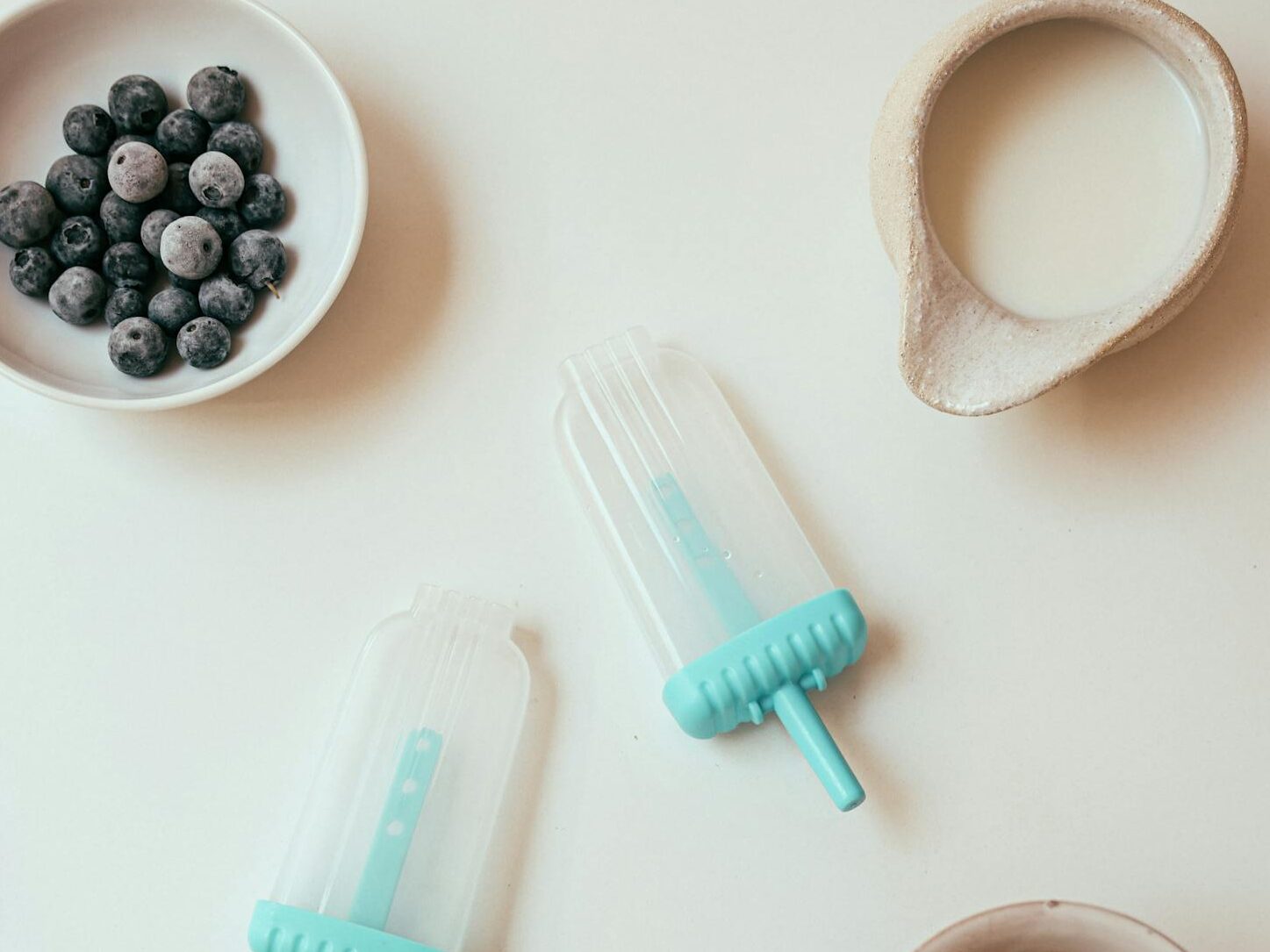 Top-down view of popsicle molds, milk, blueberries, green tea, and wooden spoon on a white surface.