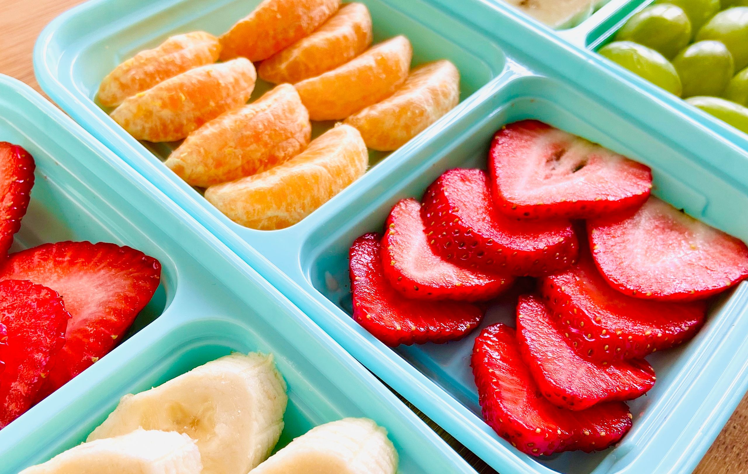 Close-up of fresh sliced fruits including oranges, strawberries, grapes, and bananas in teal trays.